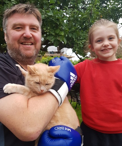 Short-haired brunette man with beard holding ginger cat and wearing blue boxing gloves, next to your girl with blonde hair tied back and wearing red jumper