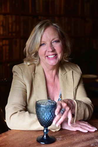 short-haired blonde woman wearing beige jacket and sitting at wooden table with blue wine glass