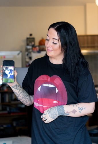 woman with long dark hair wearing black t-shirt holding up phone showing photo of tabby-and-white kitten on the screen