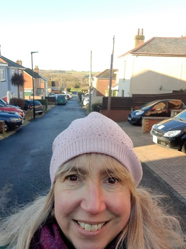 woman with long blonde hair wearing pink woolly hat with residential street behind her