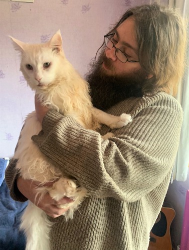 man with long brown hair and beard wearing grey jumper and holding long-haired white cat