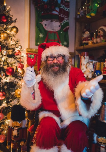 man with grey beard dressed in red and white Santa outfit and holding red Christmas bell sitting in front of Christmas tree