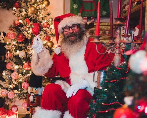 man with grey beard dressed in red and white Santa outfit and holding red Christmas bell sitting in front of Christmas tree