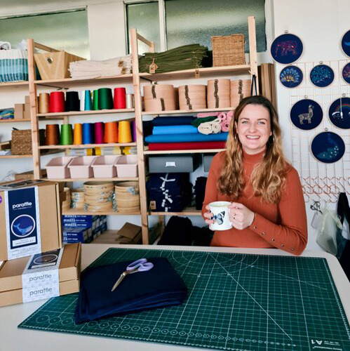 long-haired brunette woman wearing orange jumper and holding mug in a crafting studio