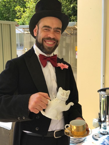 man wearing black top hat, red bowtie, and black tailcoat holding cat-shaped teapot and gold teacup