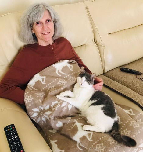 grey-haired woman sitting on cream leather sofa with grey-and-white cat with no eyes lying on a brown patterned blanket on her lap