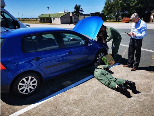 Person lying on floor under a blue car while another person looks under the bonnet