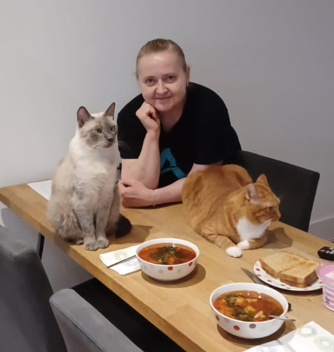 woman sitting at wooden table with one grey cat and one ginger-and-white cat sat on it alongside bowls of food