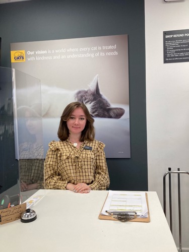 Woman with brunette hair and wearing yellow checked shirt standing behind shop counter