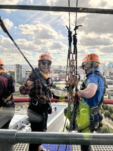 Woman wearing helmet and abseiling gear waving through window of tower