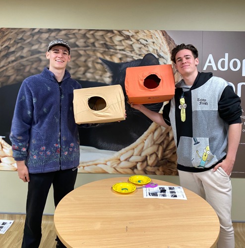 Two men holding up cat beds made from cardboard boxes and t-shirts