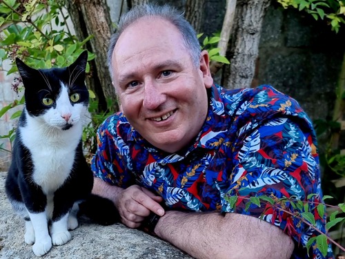 man with short grey hair and blue-and-red patterned shirt next to black-and-white cat sitting on a stone wall