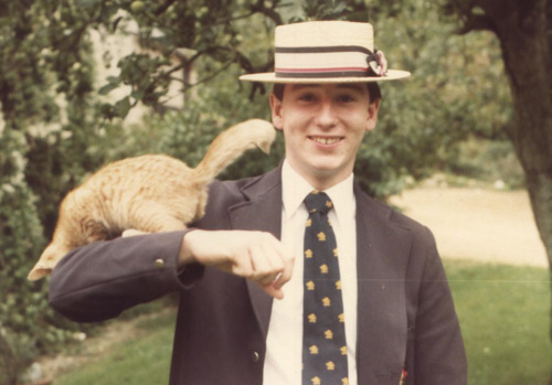 old photograph of a boy in a black blazer, white shirt, black tie and boater cat, with a ginger cat on his shoulder