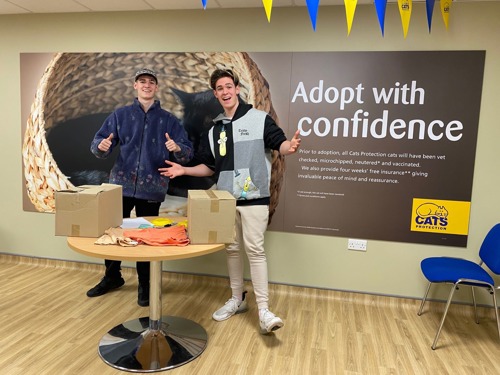 Two man standing in front of Cats Protection 'adopt with confidence' sign and behind a table with cardboard boxes and t-shirts on it