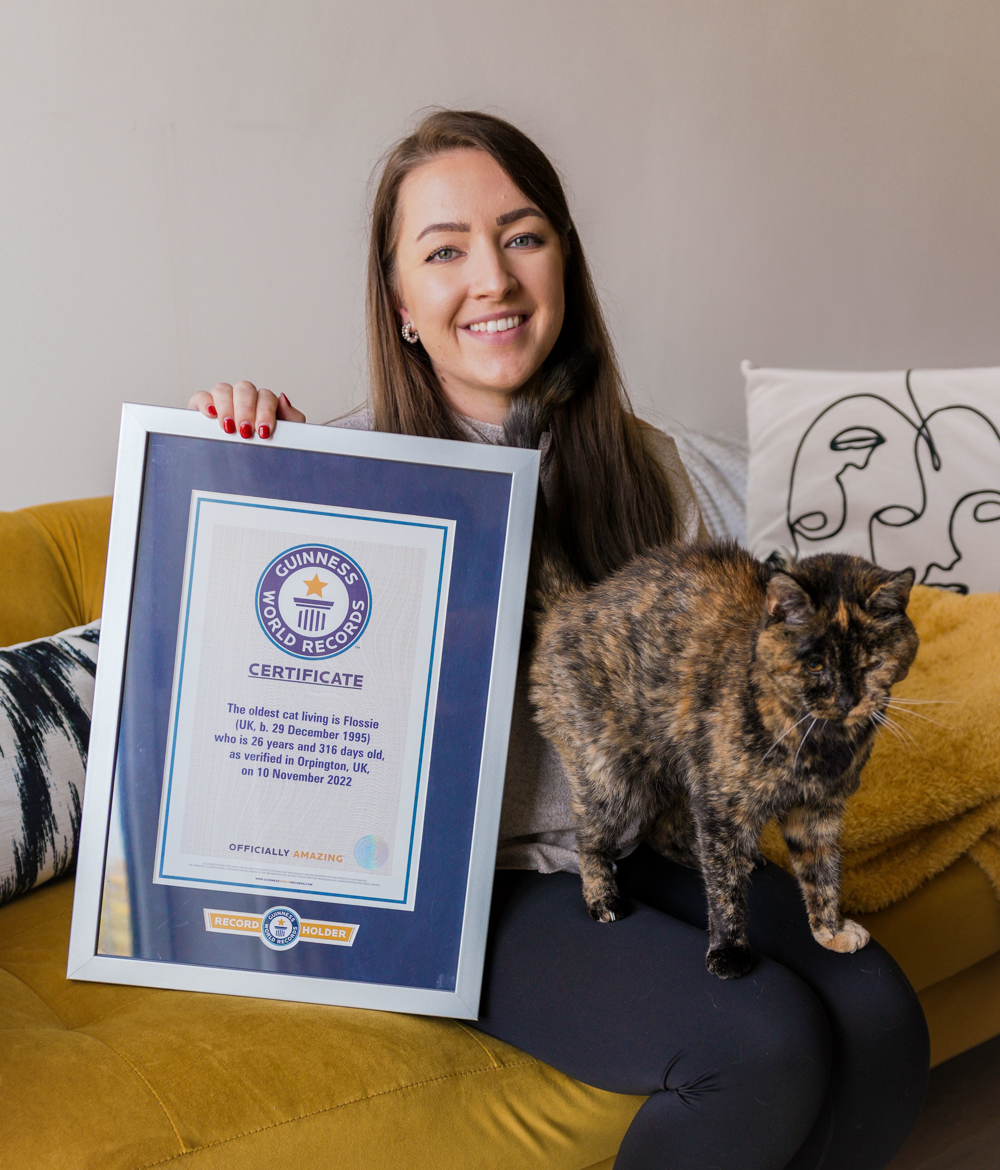 brunette woman with tortoiseshell cat on her lap and holding framed Guinness World Records certificate