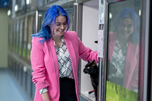 A woman with long blue hair and wearing a pink blazer stroking a black cat in a cat pen at a cat centre