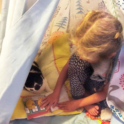 young blonde girl reading book to black and white cat inside tent