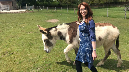 brunette woman walking in a field with a white and brown donkey