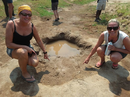 two women pointing to a lion paw in the ground in the wild