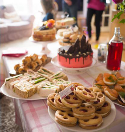 afternoon tea on a table – cakes, paw jammy biscuits, sandwiches and sausage rolls