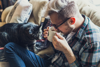 man drinking tea on sofa next to tabby cat
