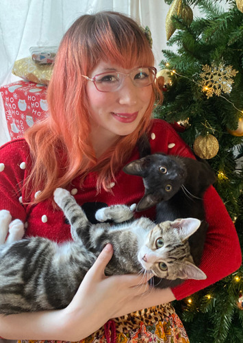 woman with pink hair holding two cats by Christmas tree
