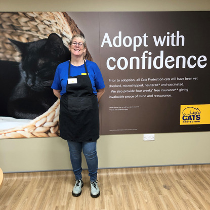 Blonde woman wearing glasses, blue Cats protection t-shirt and black apron standing in front of 'Adopt with confidence' sign
