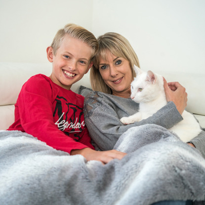 blonde boy in red top and blonde woman in grey top sitting on sofa with grey blanket and holding white cat