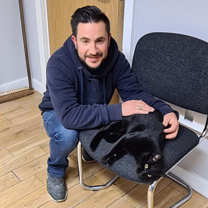 Short-haired brunette man crouched next to black cat lying on a blue chair