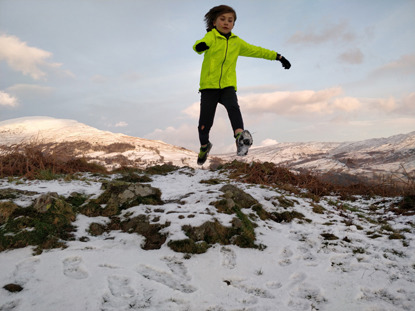 young boy wearing yellow jacket mid jump along snowy mountain path