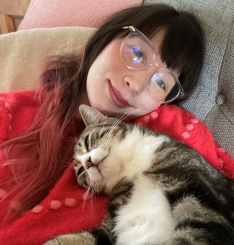 Brunette woman wearing glasses and red jumper lying on sofa with tabby-and-white cat