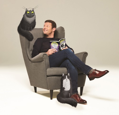 Brunette man sitting in grey armchair holding open book with illustrated grey cat sat on back of chair and illustrated black-and-white cat sat at his feet