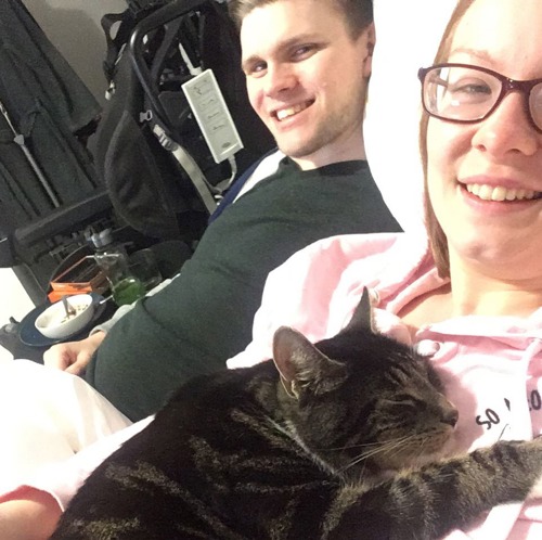 young man in black t-shirt and young woman in pink top with brown tabby-and-white cay lying on her chest