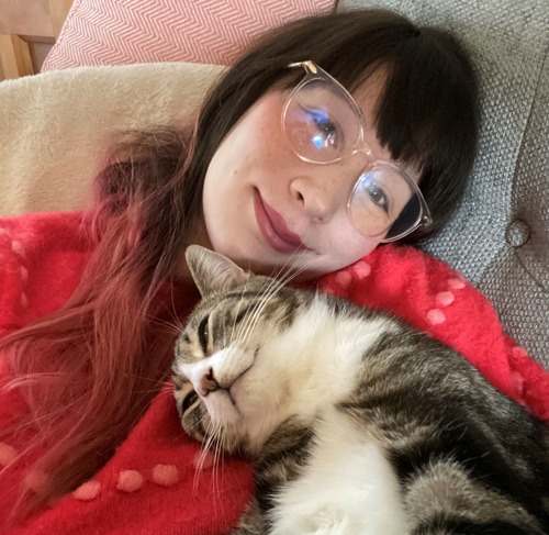 woman with long raid hair and wearing glasses cuddling tabby-and-white cat