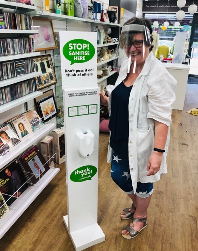 short-haired brunette woman wearing plastic face shield and white shirt standing next to hand sanitiser station in shop