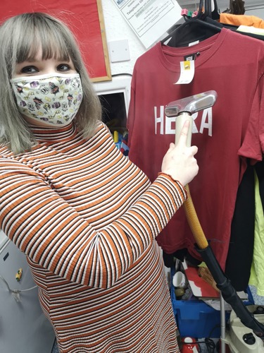 Short-haired blonde woman wearing face mask and stripy orange dress steaming a hanging red t-shirt