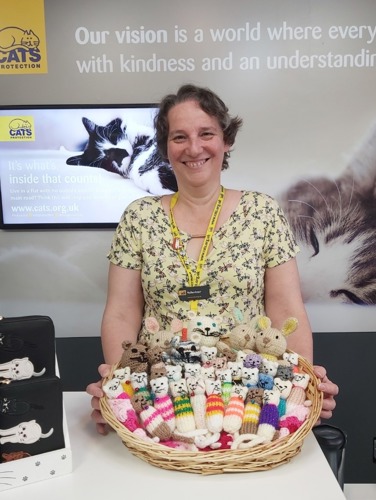 Short-haired woman standing behind Cats protection charity shop till with basket of knitted cat keyrings