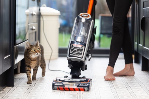tabby cat walking next to a Shark upright vacuum cleaner being pushed along by a human wearing black leggings in a kitchen