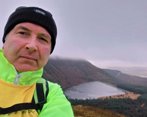 man wearing black wooly hat and high-vis jacket with mountain lake behind him