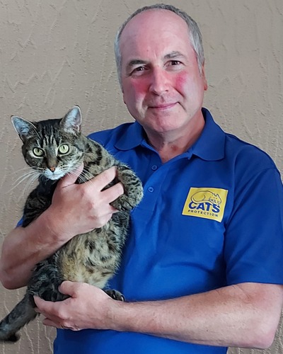 grey-haired man wearing blue Cats Protection polo shirt and holding brown tabby cat