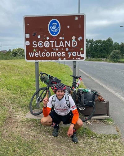 Man wearing cycling gear and helmet in front of bike and road sign saying 'Scotland welcomes you'