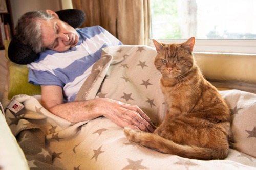 grey-haired man lying in bed with one-eyed ginger tabby cat sitting next to him