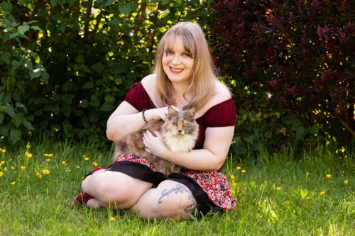 long-haired blonde woman sitting on grass holding long-haired brown-and-white cat on her lap