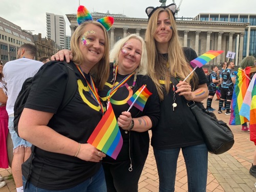 three long-haired blonde people holding rainbow flags and wearing black t-shirts with the Cats Protection Pride Network logo