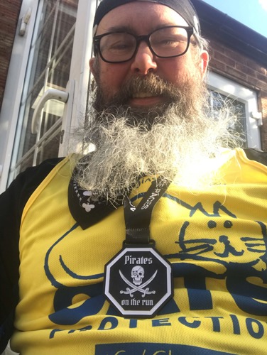 Man with long grey beard wearing yellow Cats Protection running vest and medal that says 'Pirates on the run'