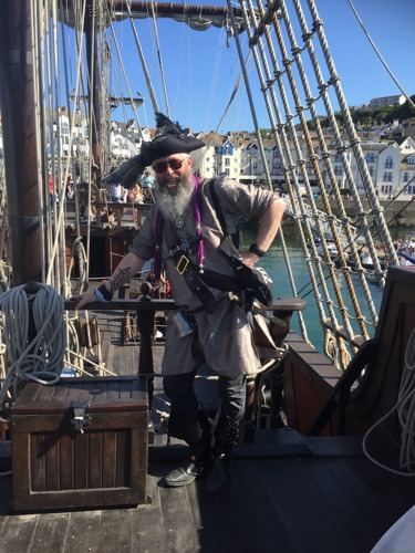 man with long grey beard dressed as a pirate standing on the deck of a ship