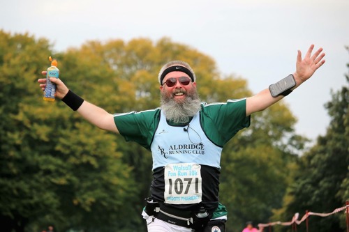 man with long-grey beard wearing running vest, crossing finish line with arms outstretched