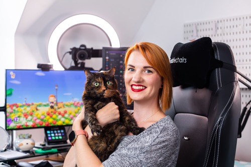 woman with short red hair sitting in black gaming chair, wearing grey top and holding tortoiseshell cat