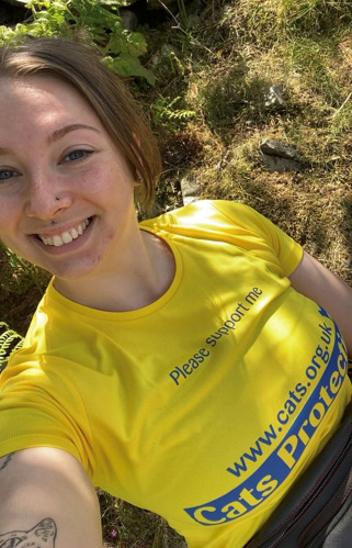 Selfie of a brunette woman wearing a yellow Cats Protection t-shirt while outdoors in front of greenery