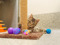 A small tabby kitten with brown and black stripes lies on the floor, playfully gripping a vertical scratching post wrapped in rope. The kitten looks directly at the camera with wide, curious eyes. Around it are several colorful toys, including purple and blue balls and a small feather toy, with an orange multi-level track toy partially visible in the background.